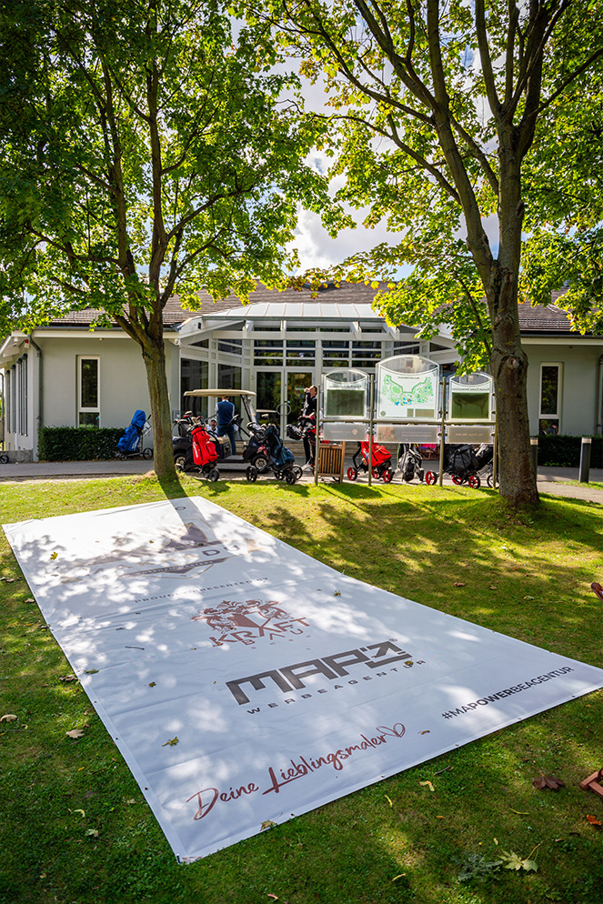 Banner mit den Logos von Makradu, Kraftbau, MAPZ Werbeagentur und dem Slogan 'Deine Lieblingsmarke' vor einem Golfclubgebäude, umgeben von Bäumen und Golfausrüstung.