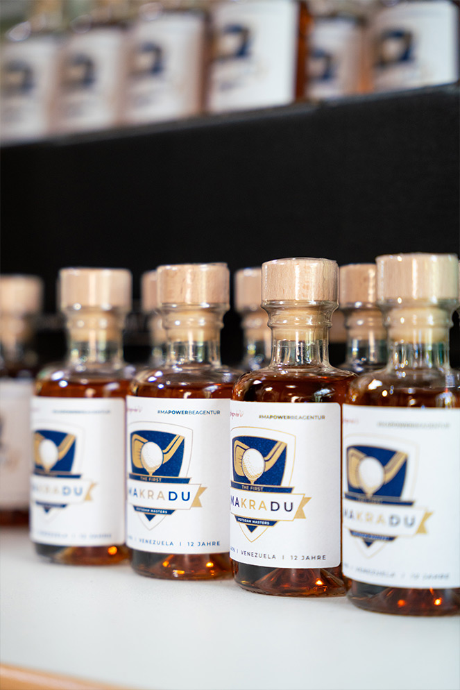 Nahaufnahme von mehreren Flaschen Makradu Rum, die nebeneinander auf einem Regal stehen. Jede Flasche hat ein Etikett mit dem Makradu-Logo und der Aufschrift 'The First Makradu', 'Venezuela', '12 Jahre'.